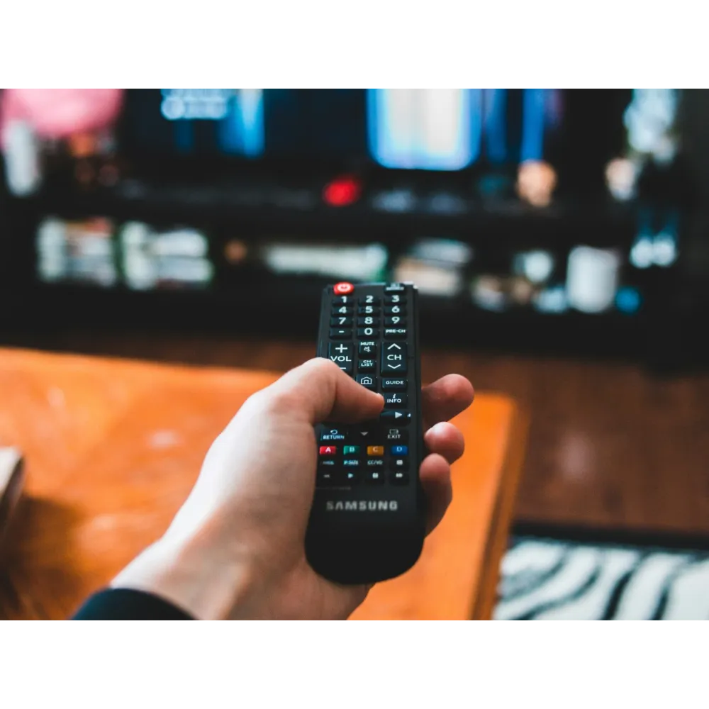 Hand holding Samsung remote control pointed at a TV screen displaying blurred content in a cozy living room.