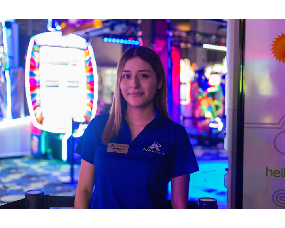 Smiling female staff member in blue Andretti uniform stands in colorful arcade with vibrant light