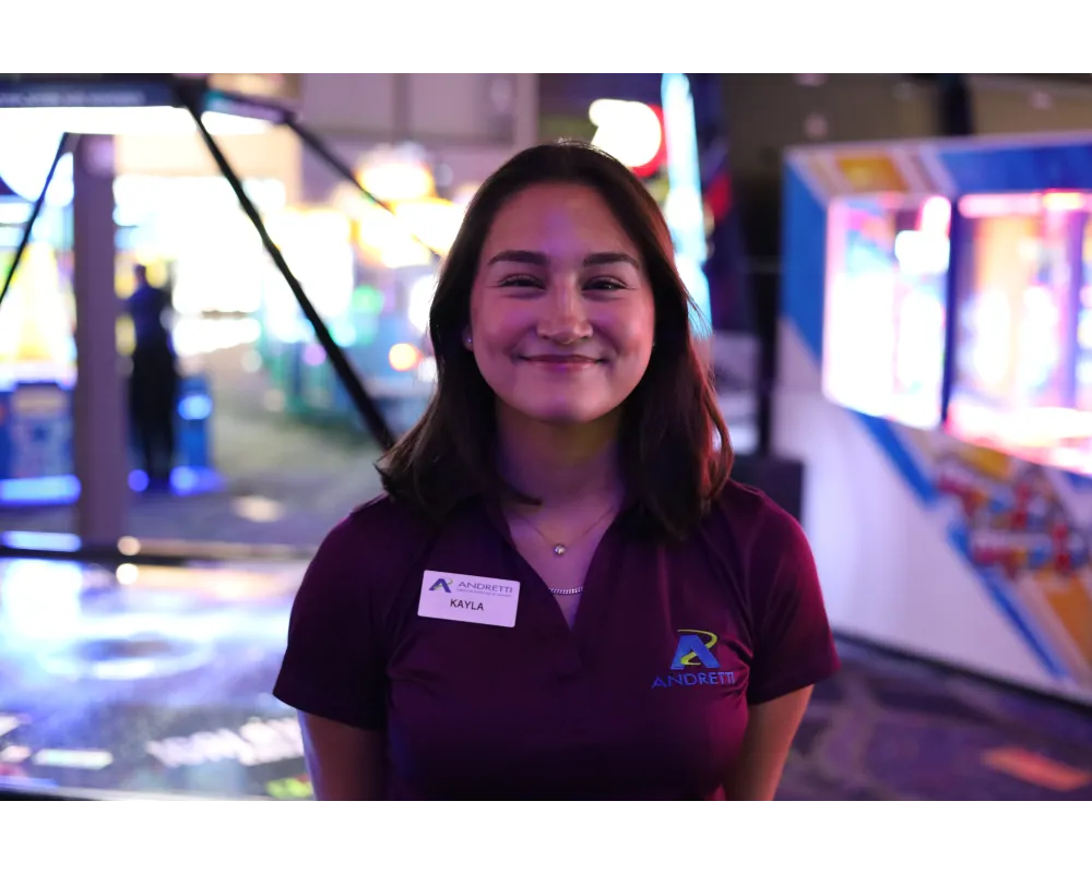 Smiling woman wearing a purple Andretti shirt and name tag standing in a colorful arcade setting.
