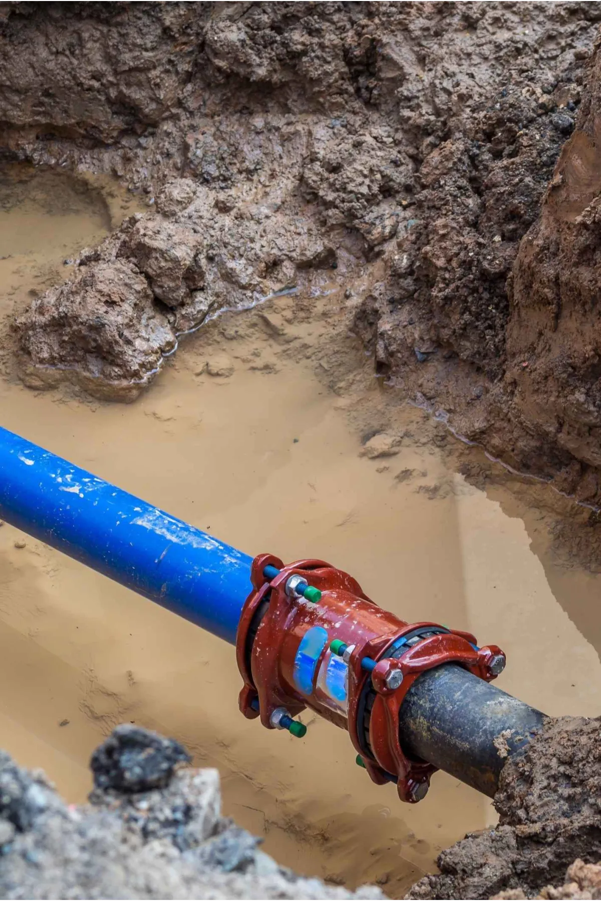 Blue and black water pipes connected by a red coupling in muddy trench with wet soil surrounding.