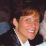 Smiling woman with short brown hair wearing a black jacket and white shirt in a dimly lit setting.