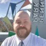 Bearded man in white shirt and tie smiling outdoors with colorful geometric sculpture and sign in background