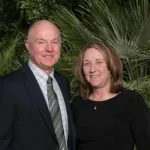 Smiling man in suit and woman in black dress posing in front of lush green tropical plants outdoors.