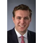 Professional headshot of a smiling man with brown hair wearing a dark suit, white shirt, and red patterned tie.