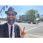 Bearded man in suit and hat making peace sign on city street with historic building and traffic lights in background