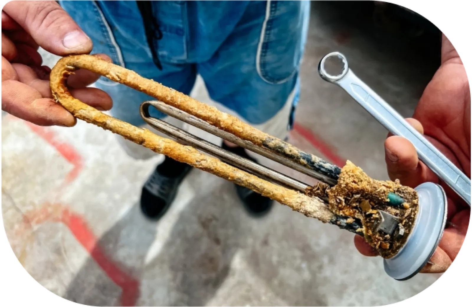 Close-up of a person's hands holding a heavily corroded water heater element and a wrench indoors.