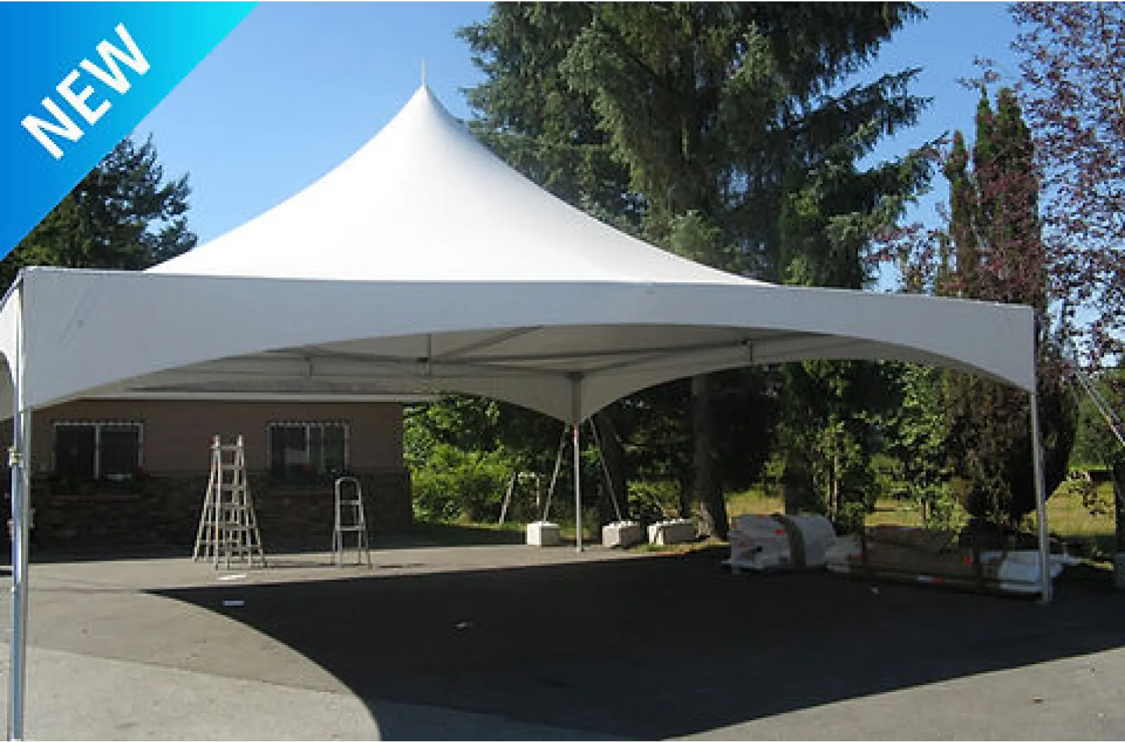 White outdoor event tent with pointed roof set up on pavement near trees and building under clear sky.