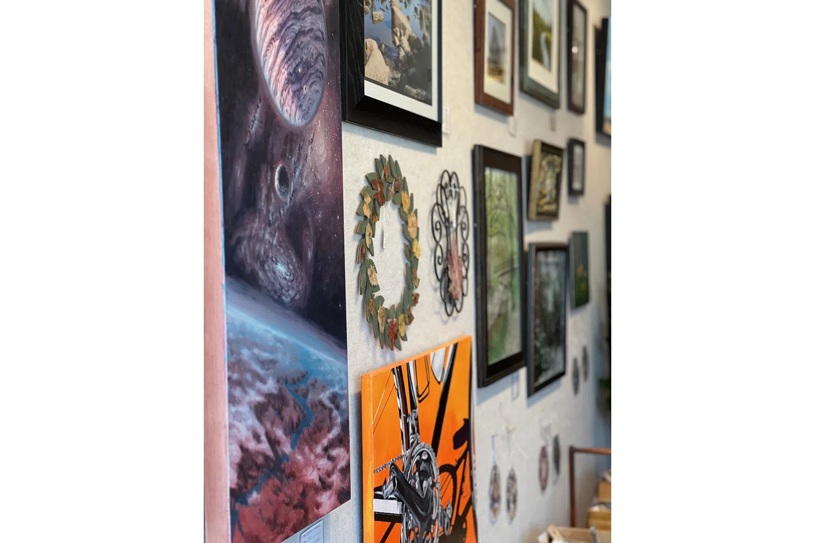 Wall display of various framed art pieces including a space scene, a bicycle painting, and decorative wreaths in a gallery.