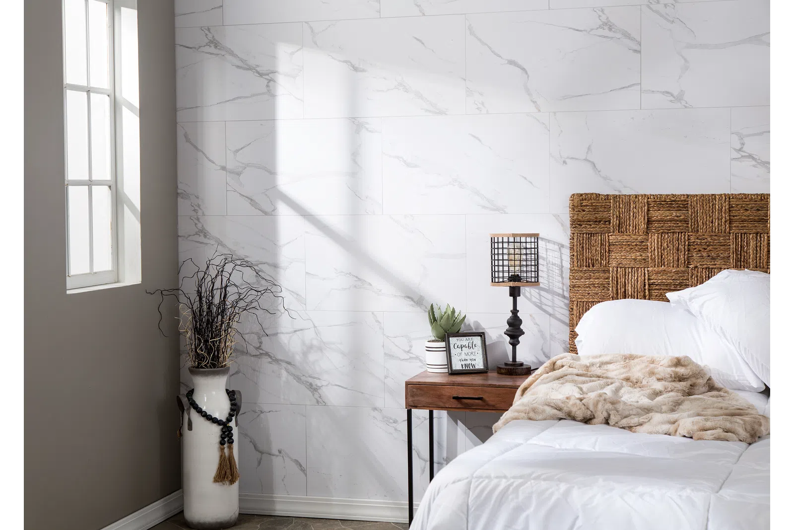 Bright bedroom corner with wicker headboard, white bedding, wooden nightstand, lamp, and decorative vase by window