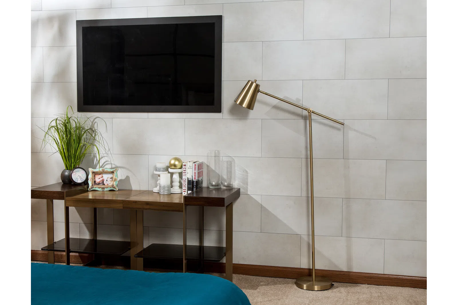 Modern bedroom corner with wall-mounted TV, wooden console table, gold floor lamp, and decorative plants and books.