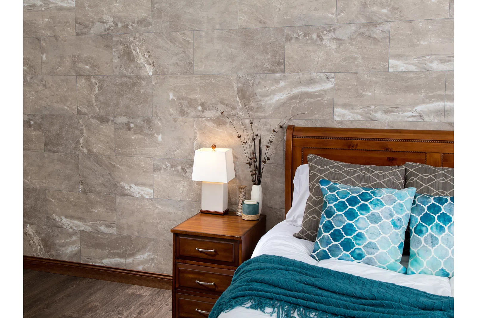 Cozy bedroom corner with wooden bed, blue patterned pillows, teal throw, nightstand, and table lamp on marble wall.
