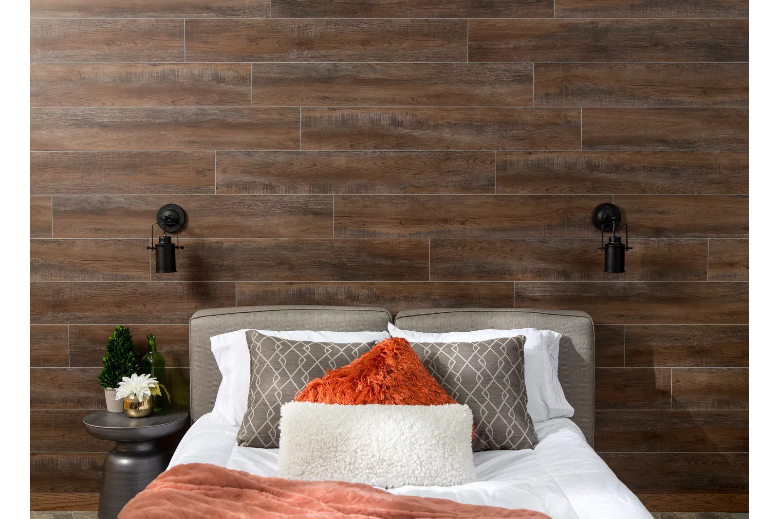 Cozy bed with textured pillows and throw against a dark wooden plank accent wall with wall-mounted lamps.