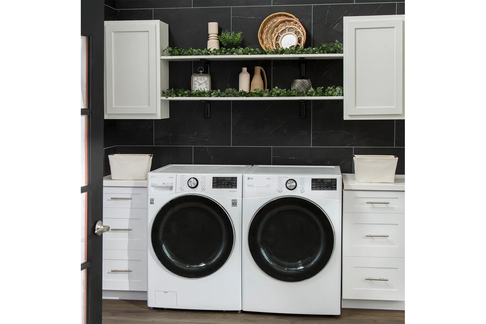 Modern laundry room with white front-load washer and dryer, black tiled wall, white cabinets, and decorative shelves with plants.