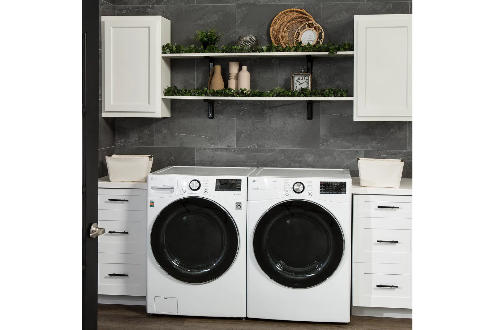 Modern laundry room with white front-load washer and dryer, white cabinets, shelves with decor, and gray tiled wall.