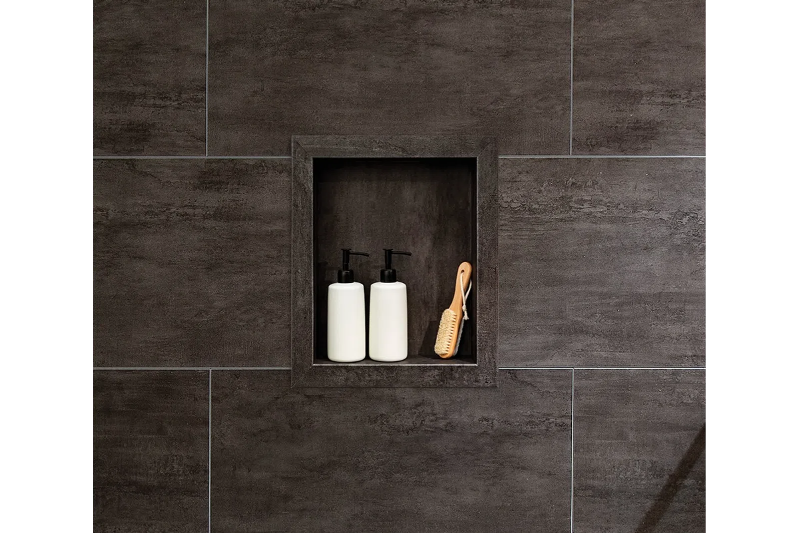 Dark tiled shower niche with two white pump bottles and a wooden bath brush neatly arranged inside