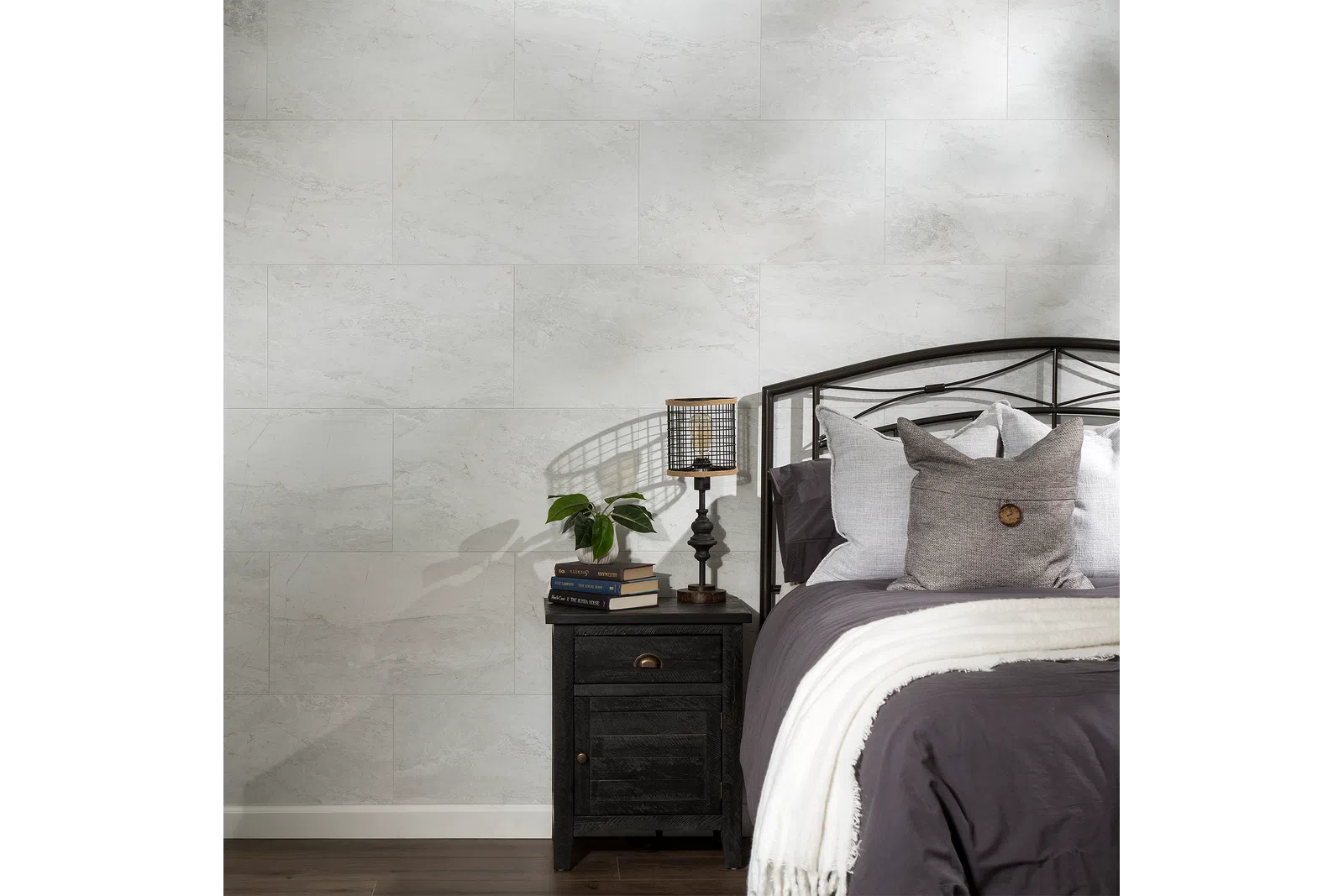 Cozy bedroom corner with dark metal bed, gray bedding, black nightstand, lamp, books, and plant against light stone wall