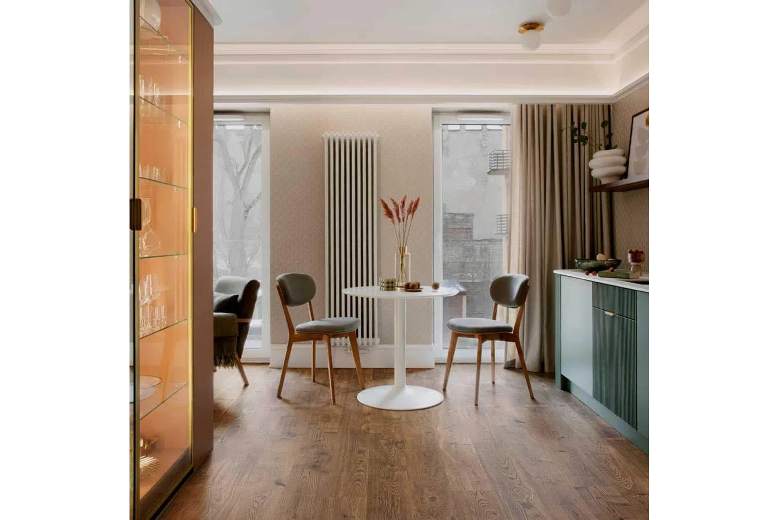 Modern dining area with round white table, two wooden chairs, cozy lighting, and green kitchen cabinets.