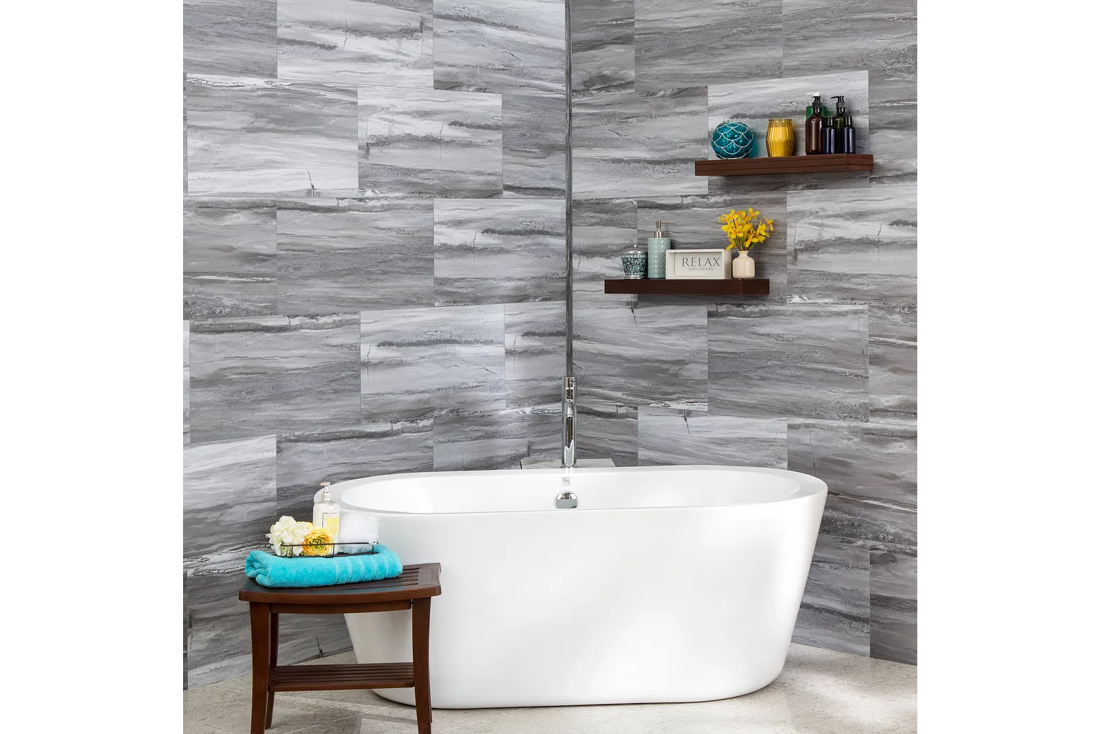 Modern white freestanding bathtub with gray stone tile walls and decorative shelves in bathroom corner.