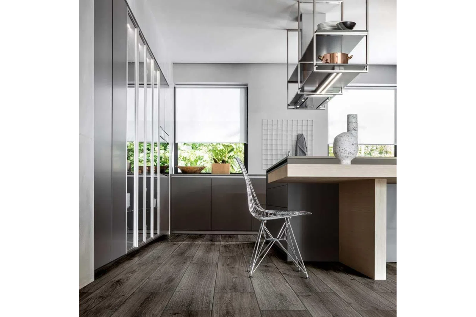 Modern kitchen with sleek gray cabinets, wooden flooring, metal wire chair, and minimalist decor under natural light.