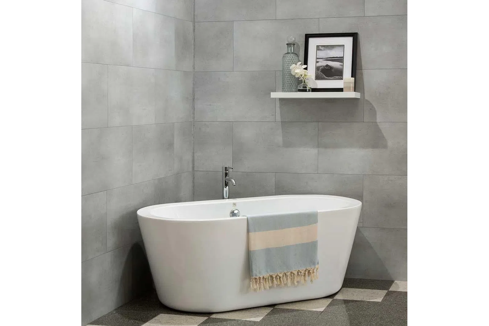 Modern white freestanding bathtub with a striped towel in a gray tiled bathroom with a floating shelf above.