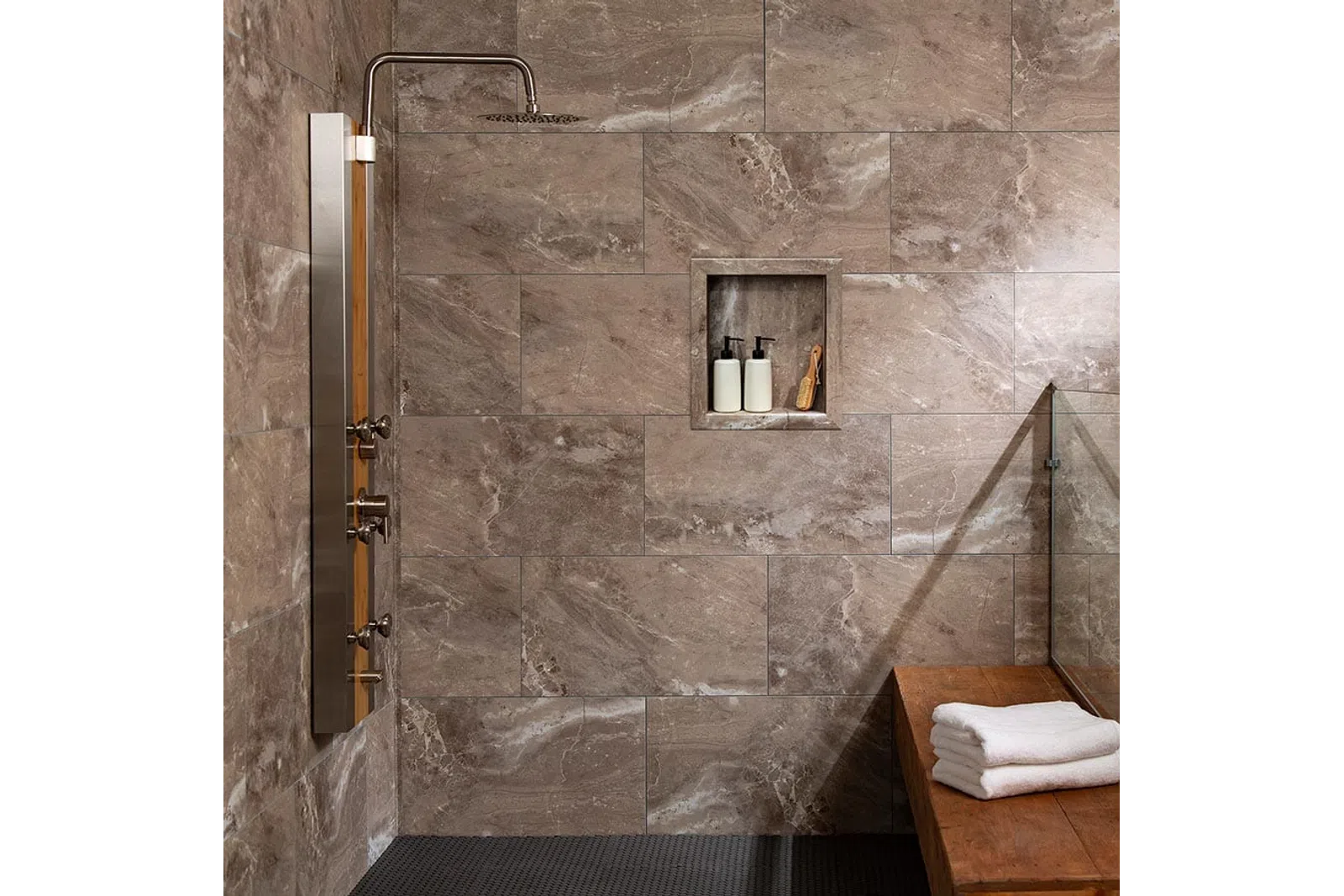 Modern shower area with large stone tiles, rain showerhead, built-in shelf with toiletries, and wooden bench with towels.