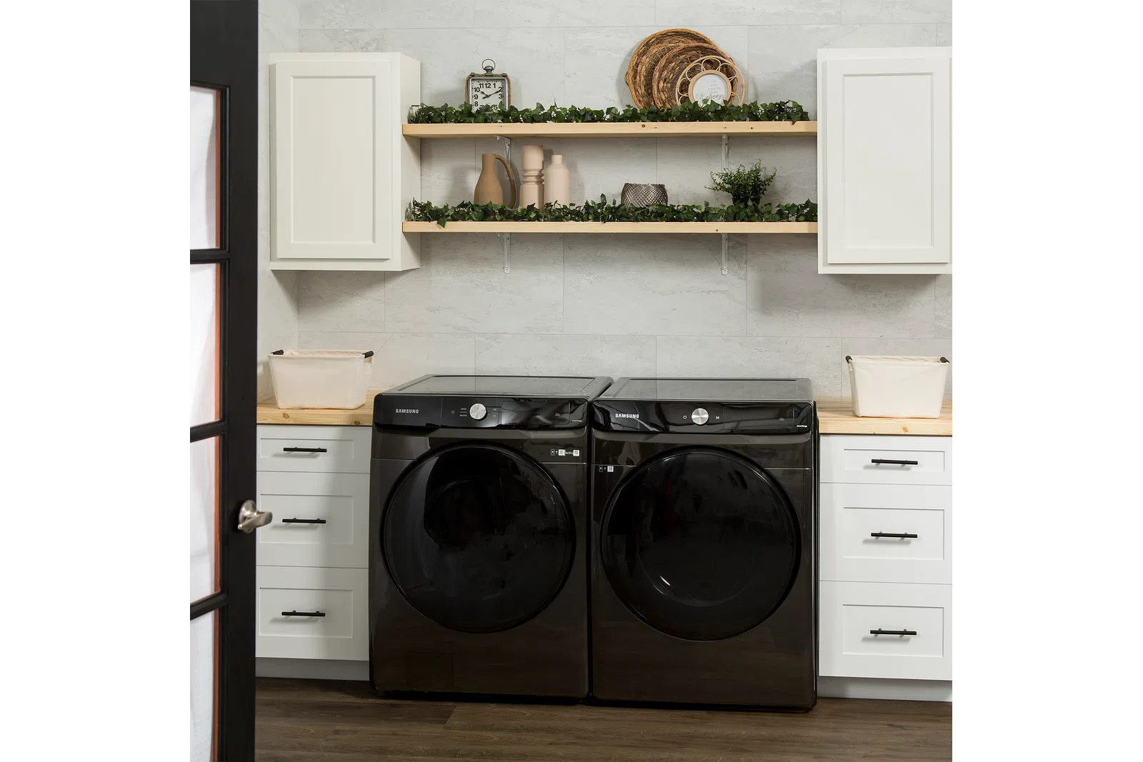 Modern laundry room with black Samsung washer and dryer, white cabinets, wooden shelves, and decorative greenery.