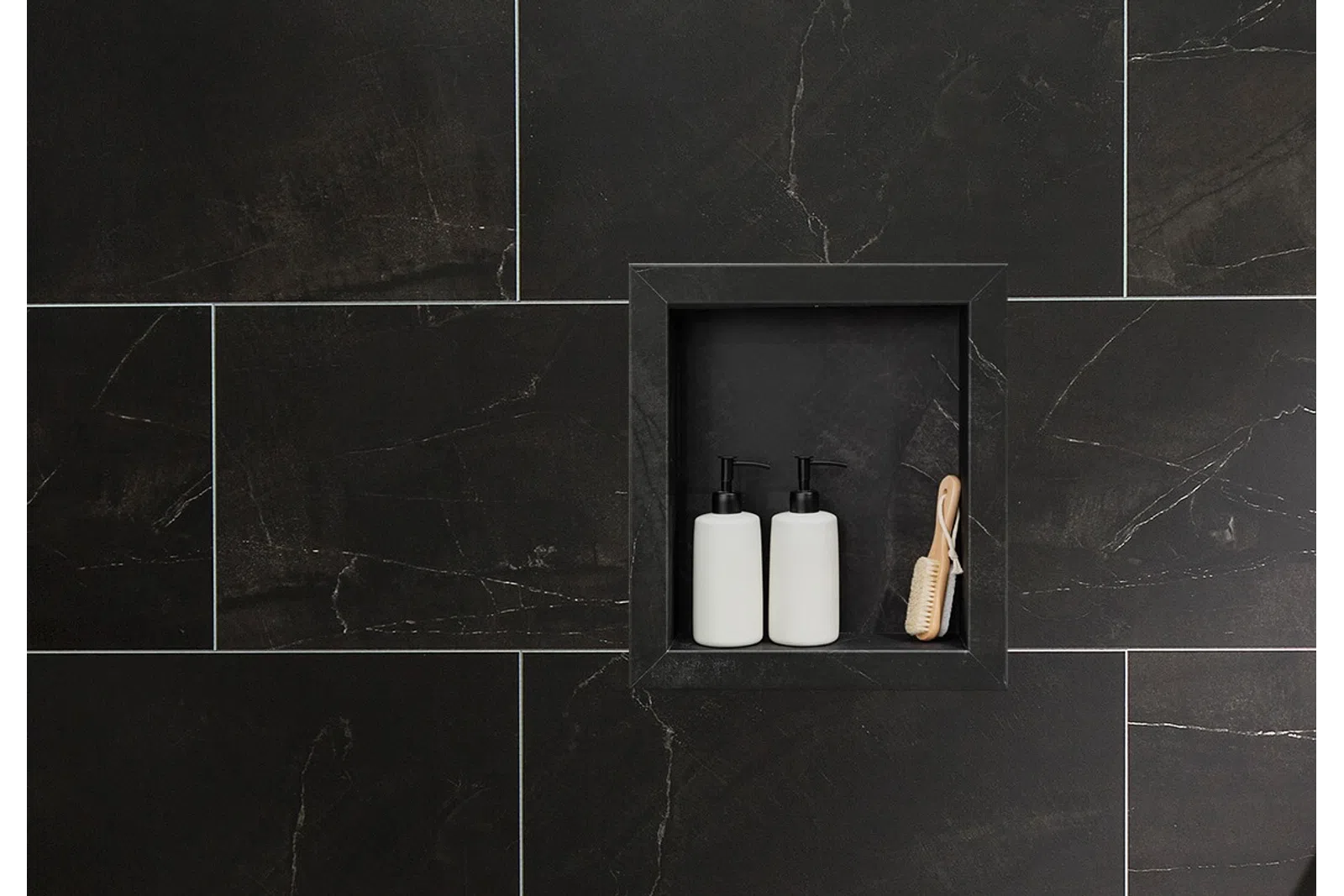 Two white pump bottles and a wooden brush in a black tiled shower niche with dark grout lines.