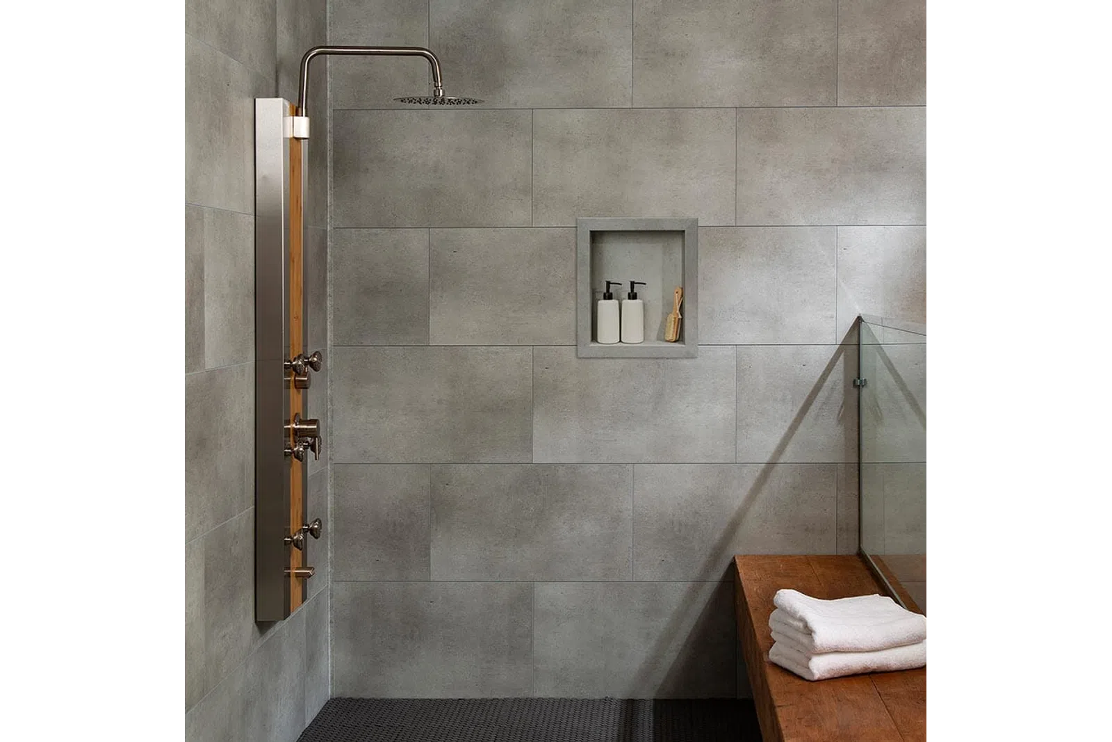 Modern shower area with gray tiled walls, built-in soap niche, wooden bench, and folded towels on bench.