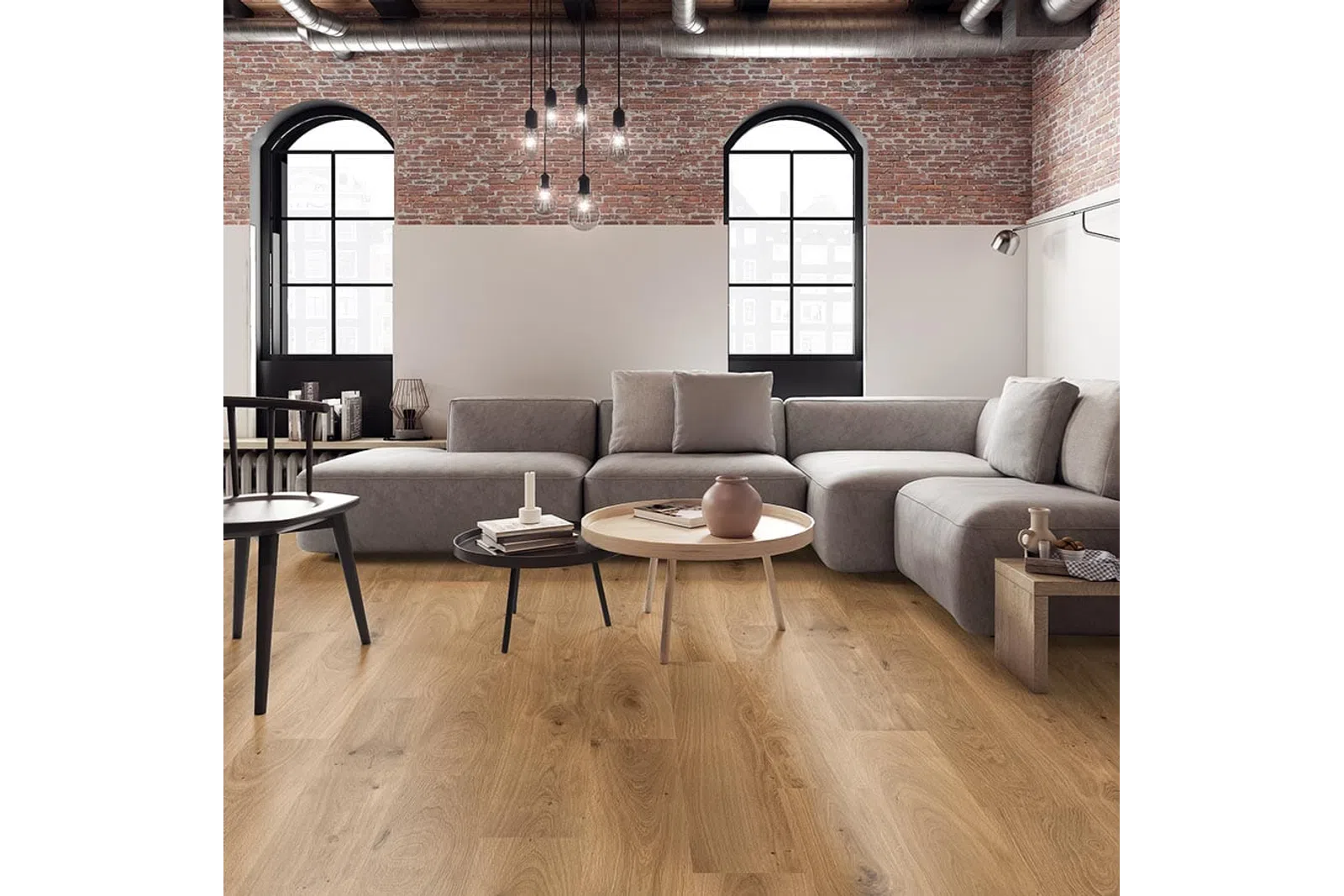 Modern living room with gray sectional sofa, wooden coffee tables, exposed brick wall, and large arched windows.