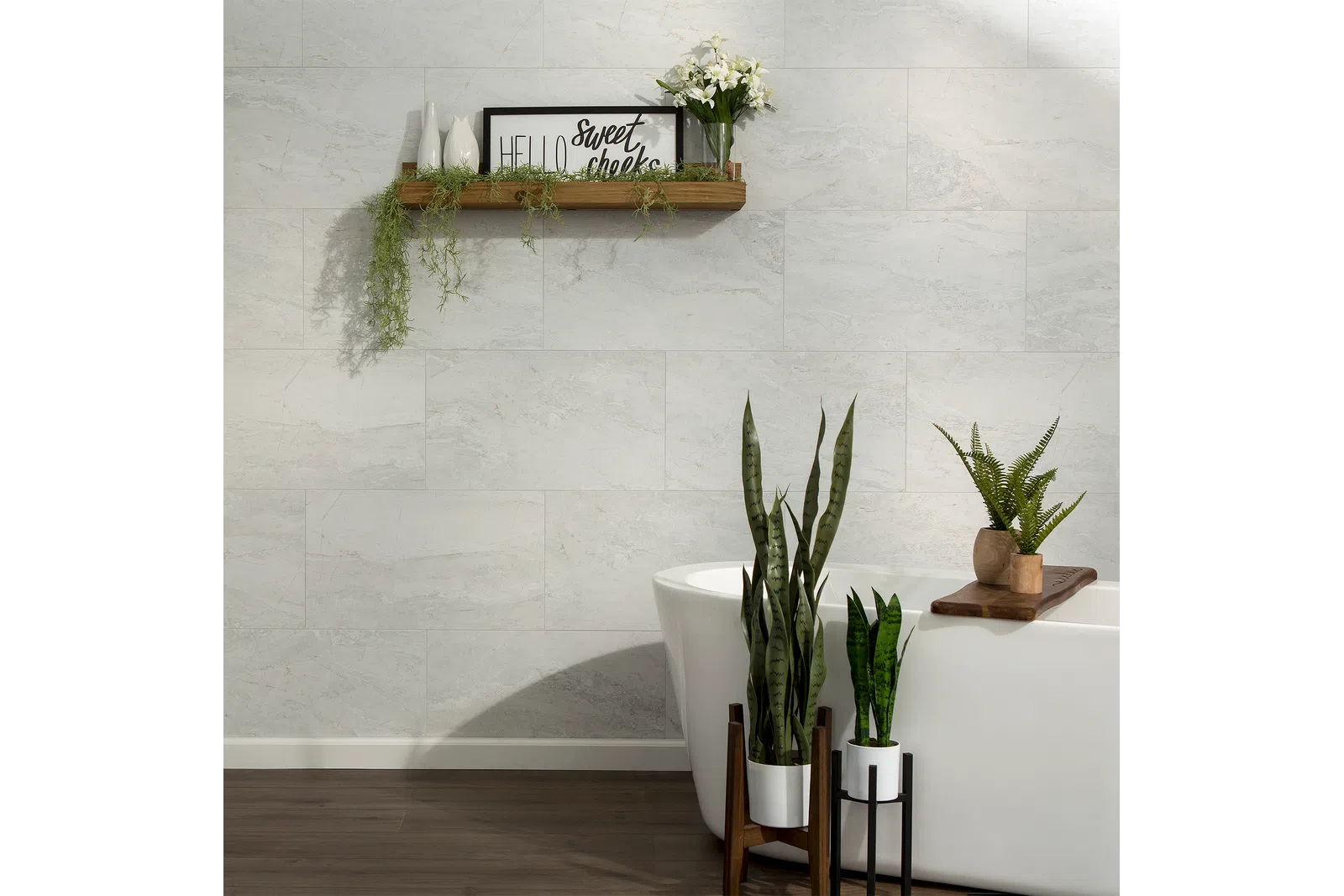 Modern bathroom with freestanding white tub, green plants, floating wooden shelf, and light gray tiled wall.