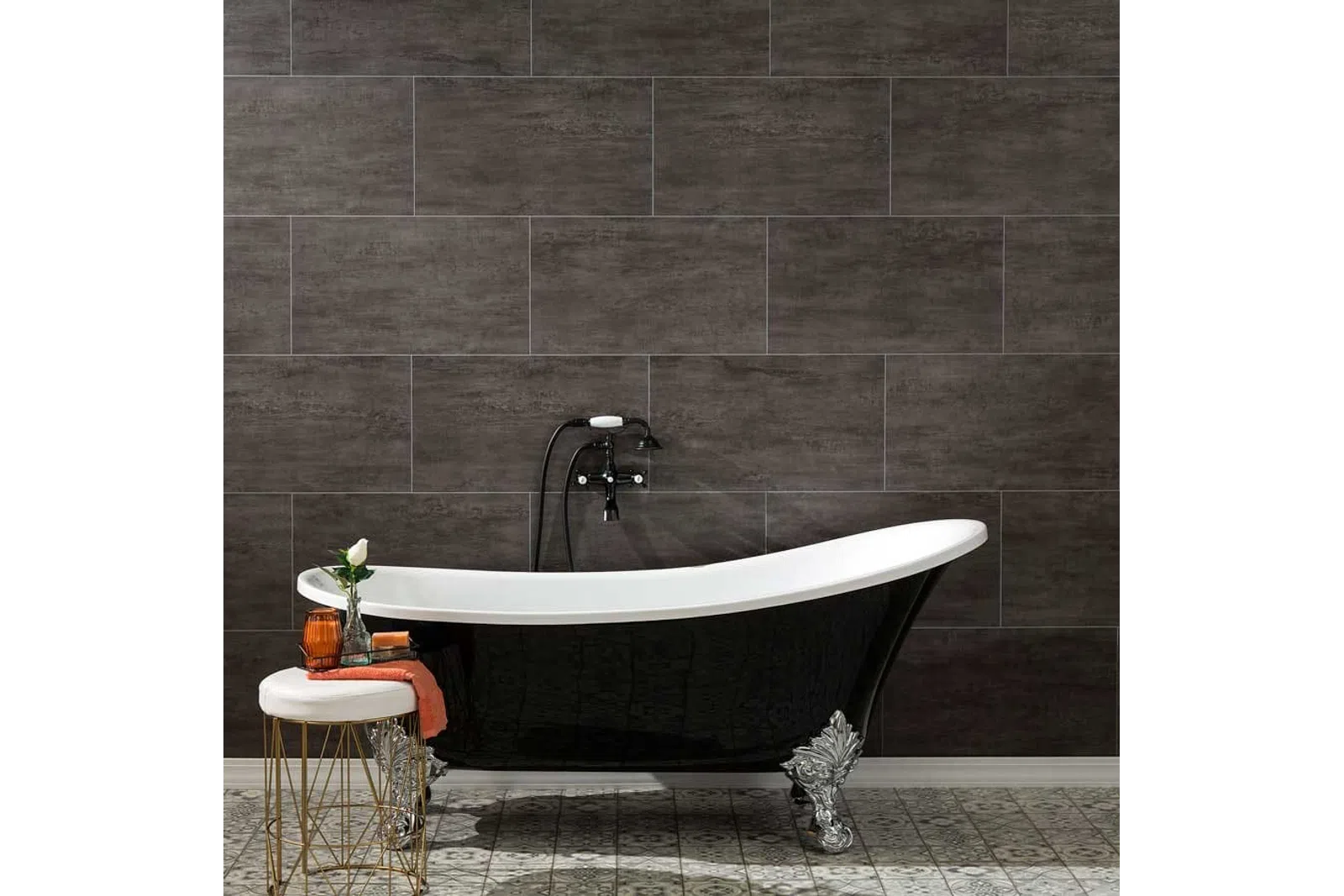 Black clawfoot bathtub with silver feet against dark tile wall and decorative stool with orange towel and vase