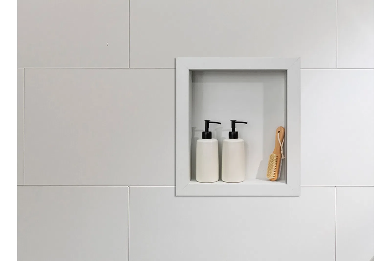 White tiled bathroom wall with recessed shelf holding two white pump bottles and a wooden brush.