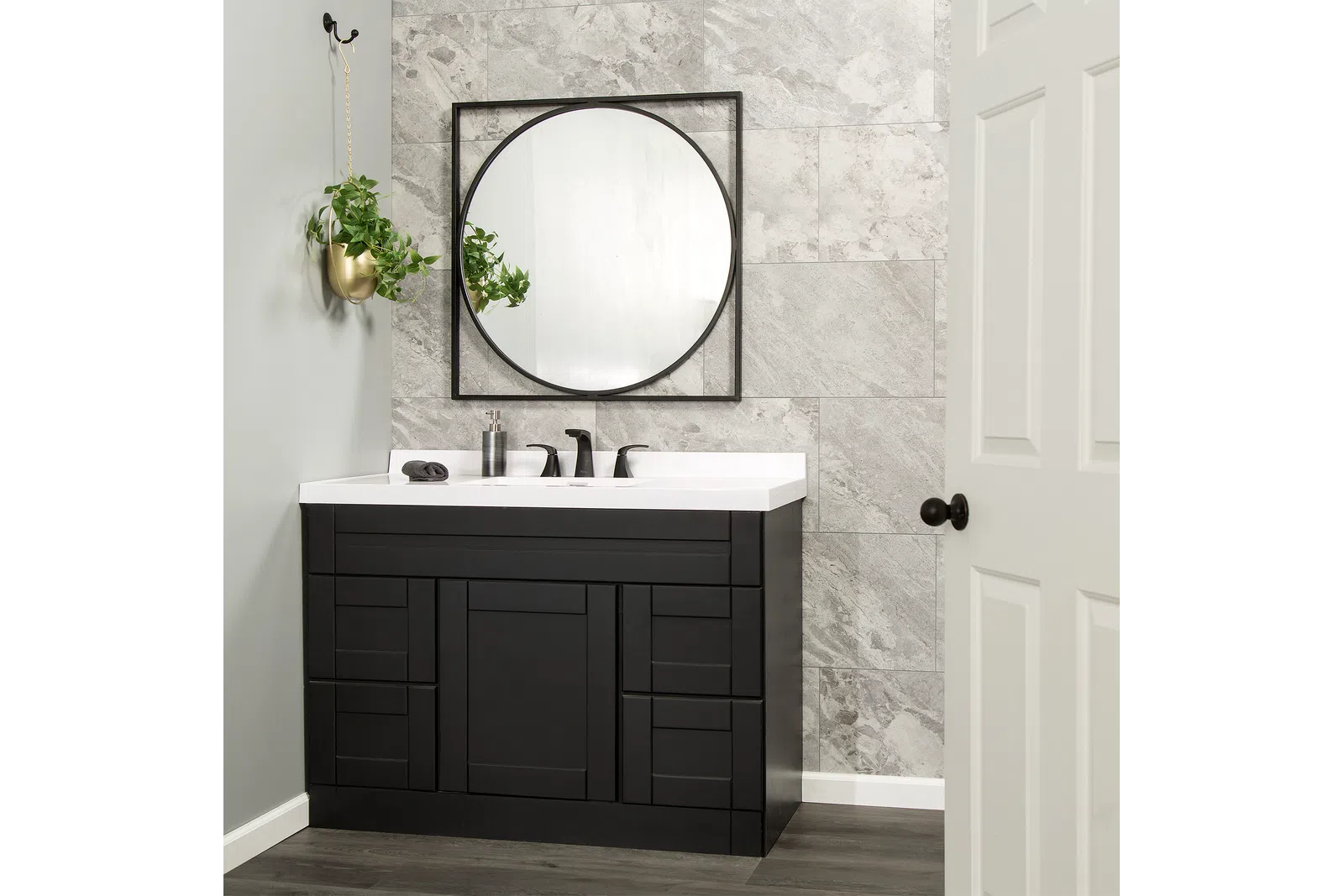 Modern black bathroom vanity with white countertop, round mirror, gray tiled wall, and hanging plant in gold pot.