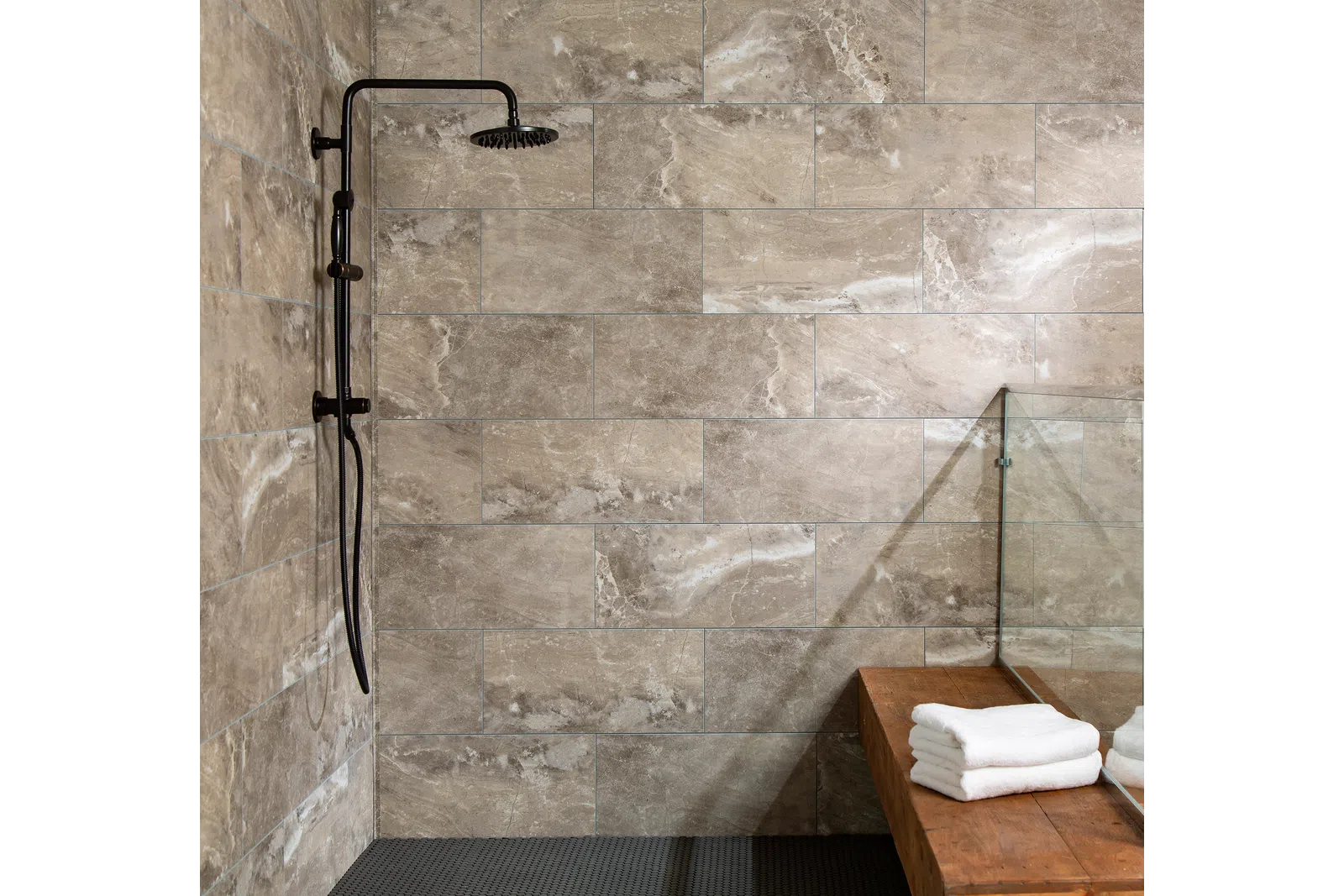 Modern bathroom shower area with beige stone tiles, black rainfall showerhead, and wooden bench with white towels.