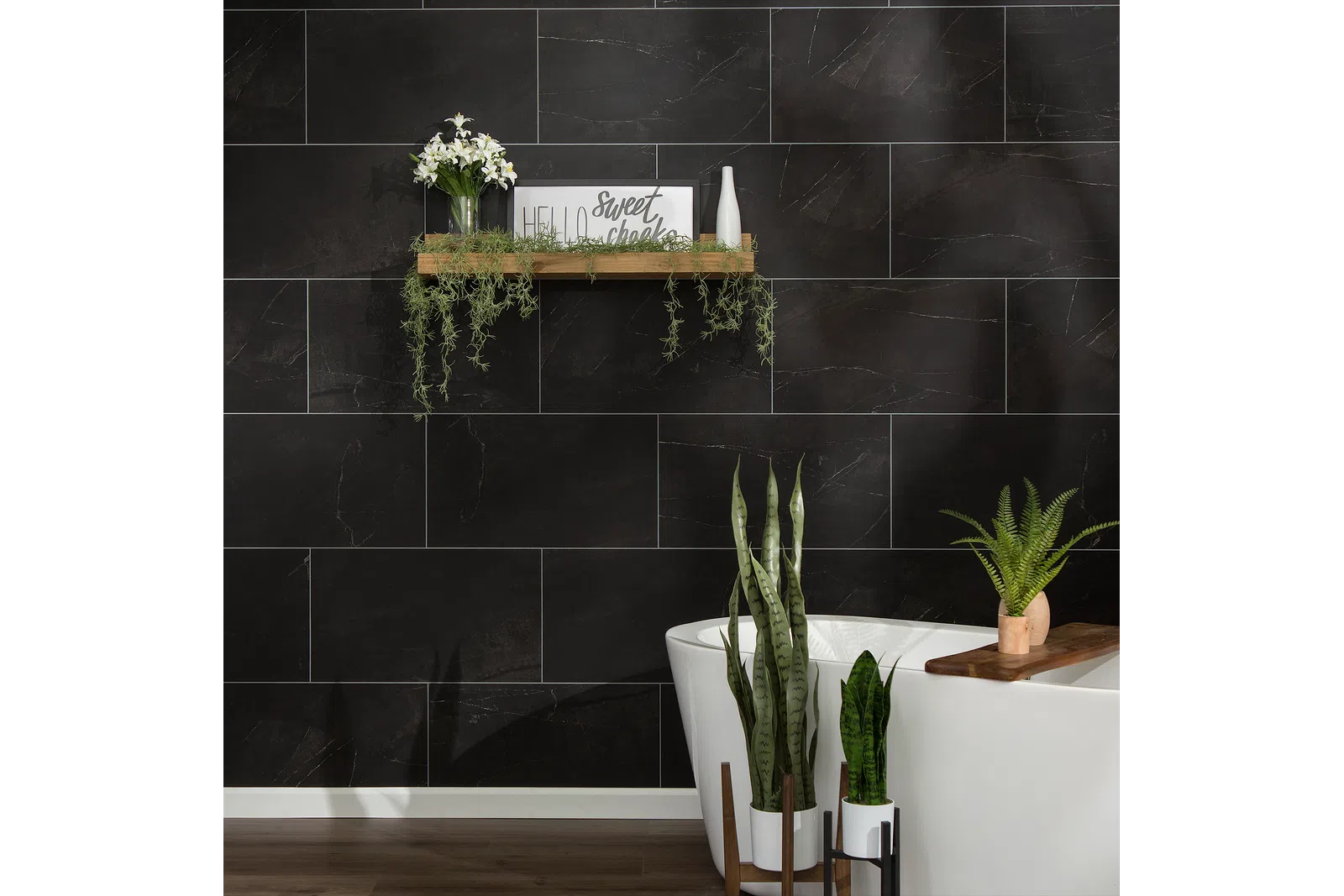 Modern bathroom with black marble tiles, white freestanding tub, wooden shelf with plants and decor.