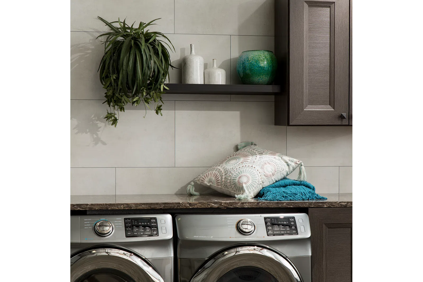 Modern laundry room with stainless steel washer and dryer, decorative pillows, hanging plant, and ceramic vases.