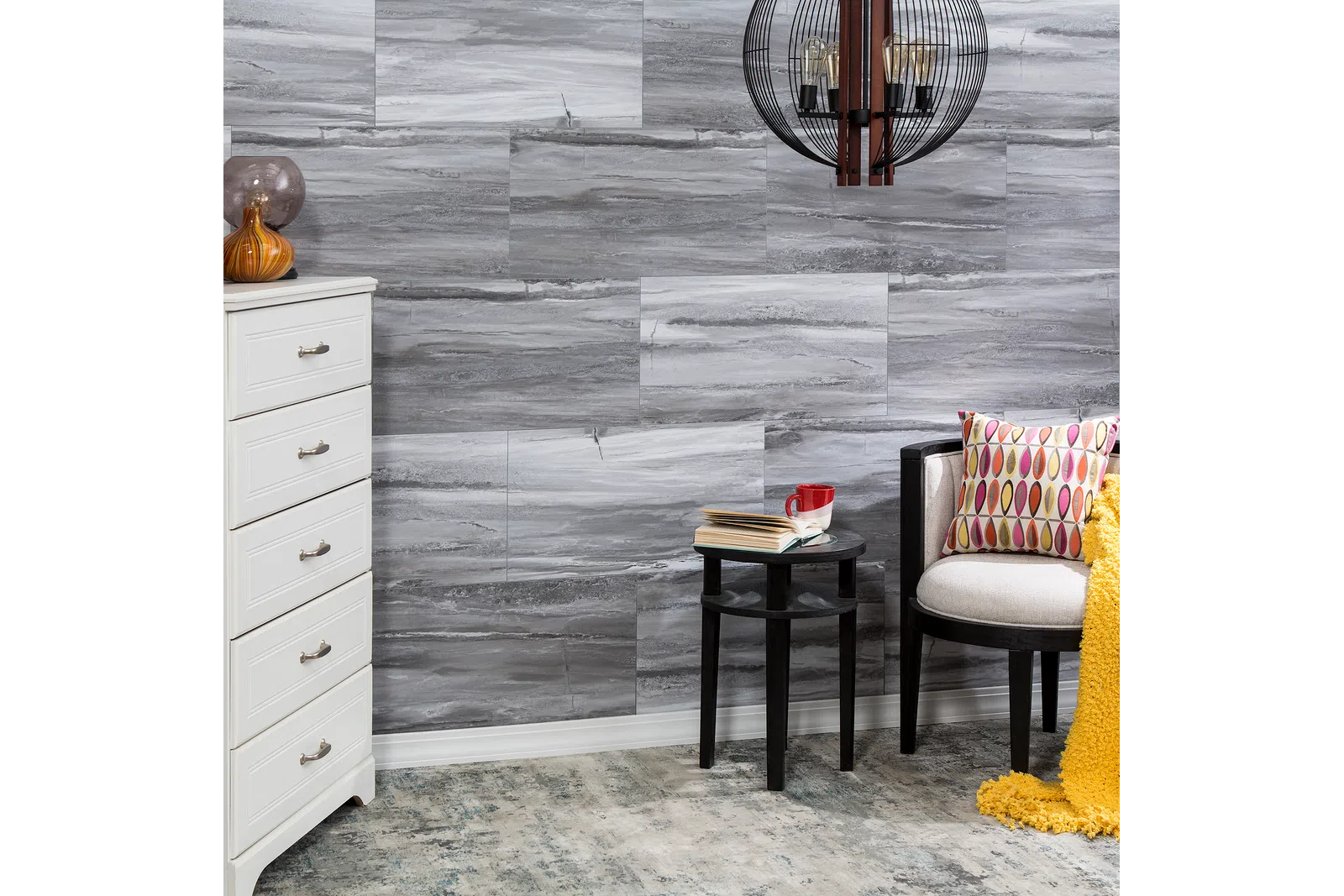 Cozy corner with white dresser, patterned pillow chair, black side table, gray stone wall, and decorative lighting fixture.
