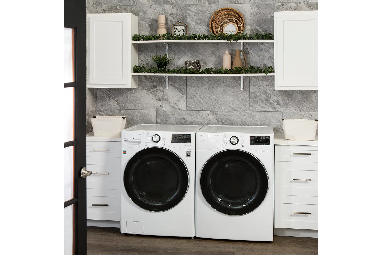 Modern laundry room with front-load washer and dryer, white cabinetry, grey stone wall, and decorative shelves.
