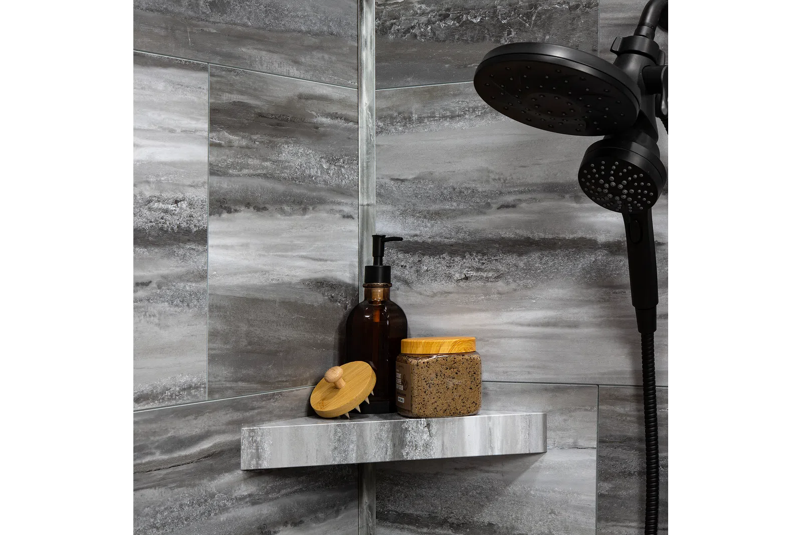 Modern bathroom shower with dark showerhead, gray marble tiles, soap dispenser, scrub brush, and exfoliating scrub jar.