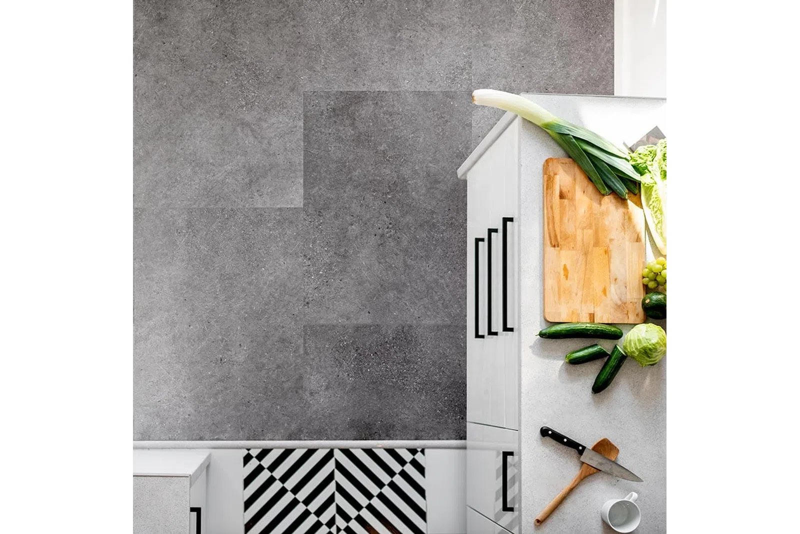 Top view of a white kitchen counter with chopping board, vegetables, knife, and modern gray tile flooring