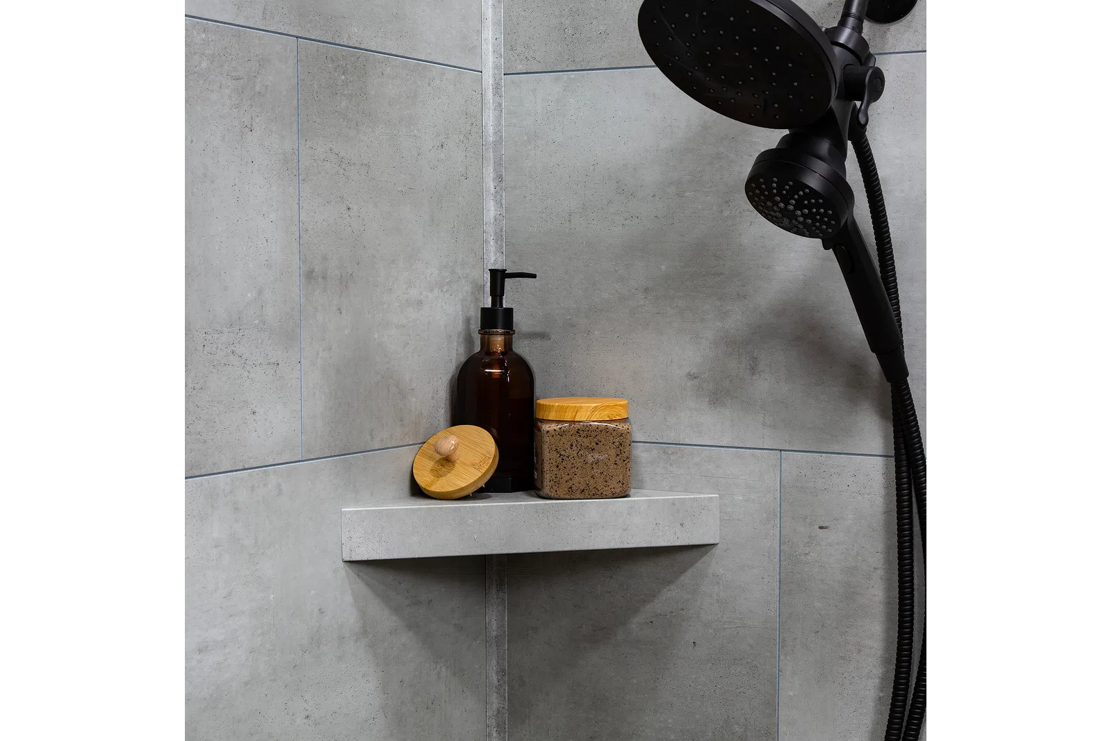 Modern shower corner with black showerhead and natural skincare containers on grey stone shelf.