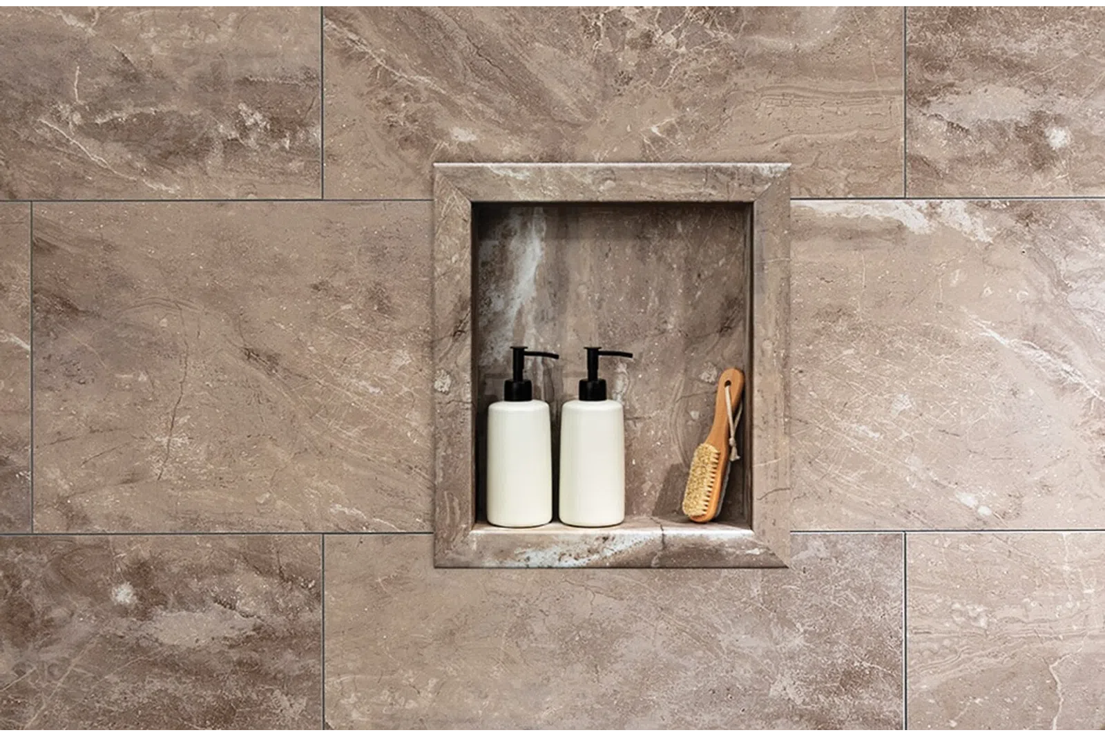 Two white pump bottles and a wooden brush placed in a stone wall niche with beige marble tiles around.