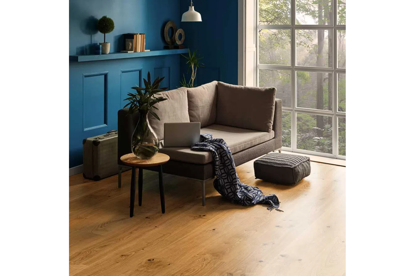 Cozy modern living room with beige sofa, laptop, blue wall, large window showing green trees outside.