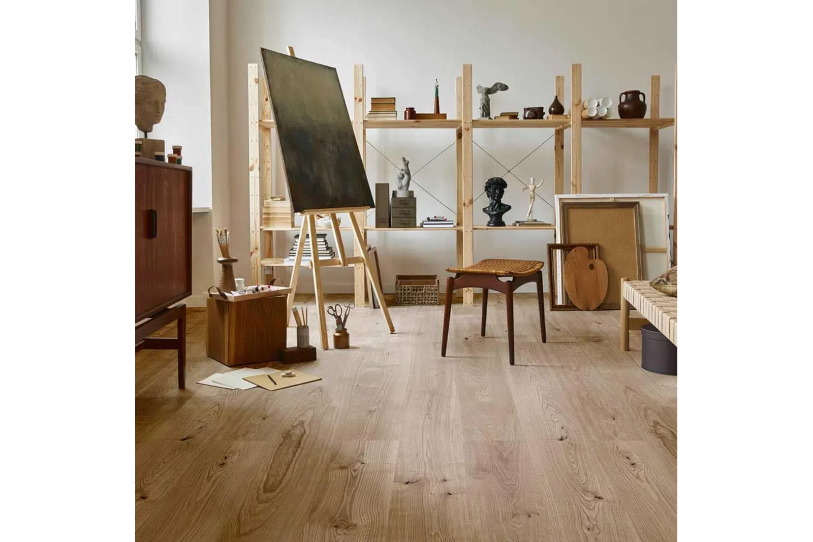 Cozy art studio with wooden flooring, easel, sculptures, wooden chair, and open shelves filled with art supplies.