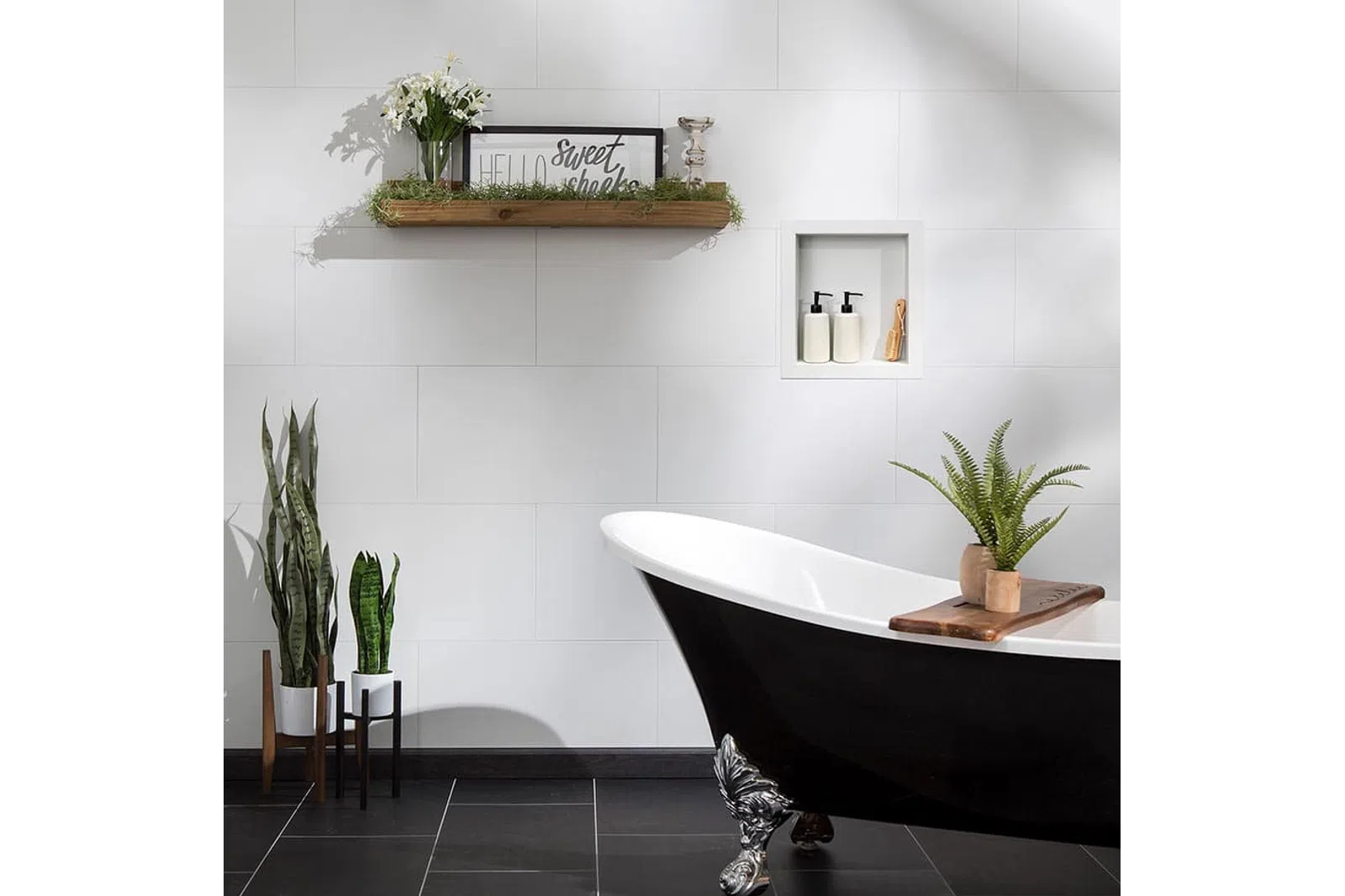 Modern bathroom with black clawfoot bathtub, white tiled walls, plants, and decorative shelf with framed sign.