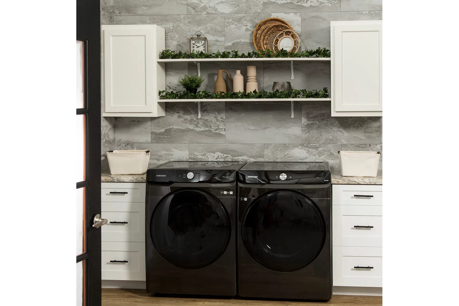 Modern laundry room with black Samsung washer and dryer, white cabinets, marble backsplash, and decorative shelves.