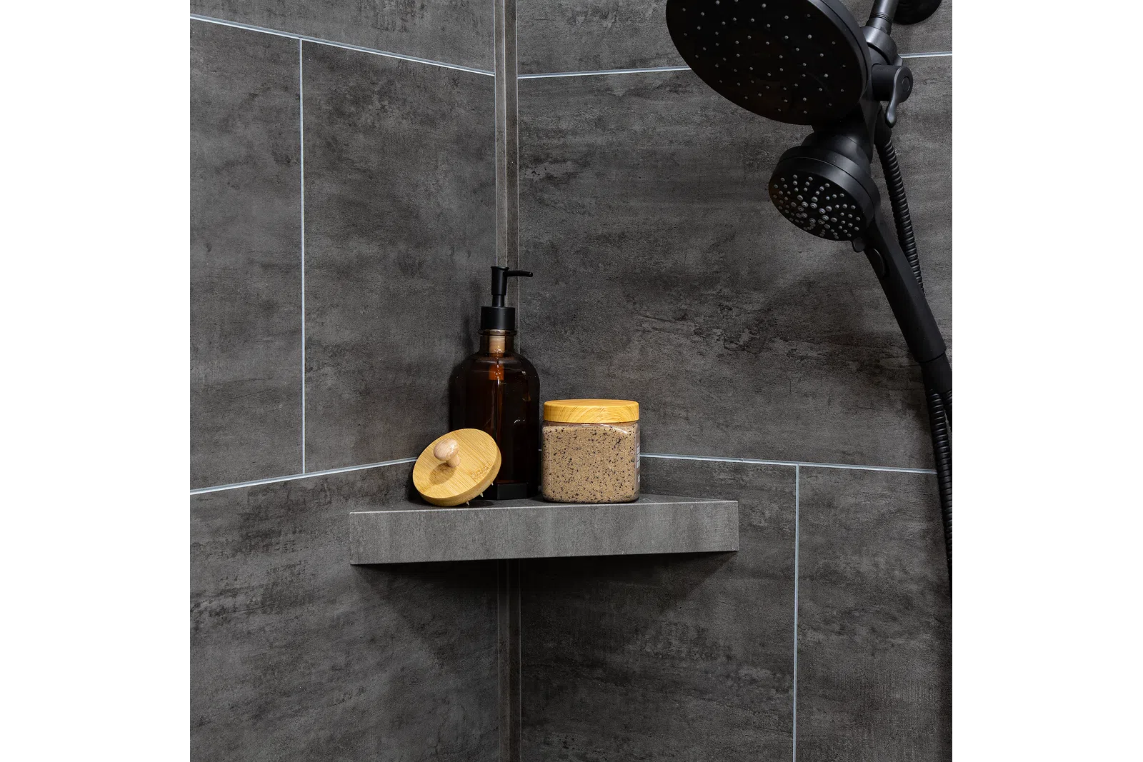 Modern shower corner with dark gray tiles, black showerhead, amber soap dispenser, and scrub jar with wooden lids.