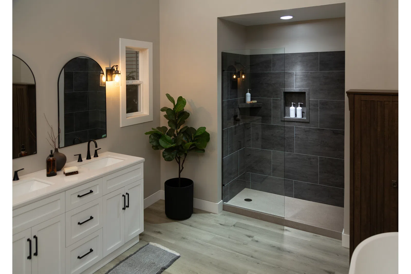 Modern bathroom with white double vanity, black fixtures, indoor plant, and walk-in shower with dark tiles and glass door