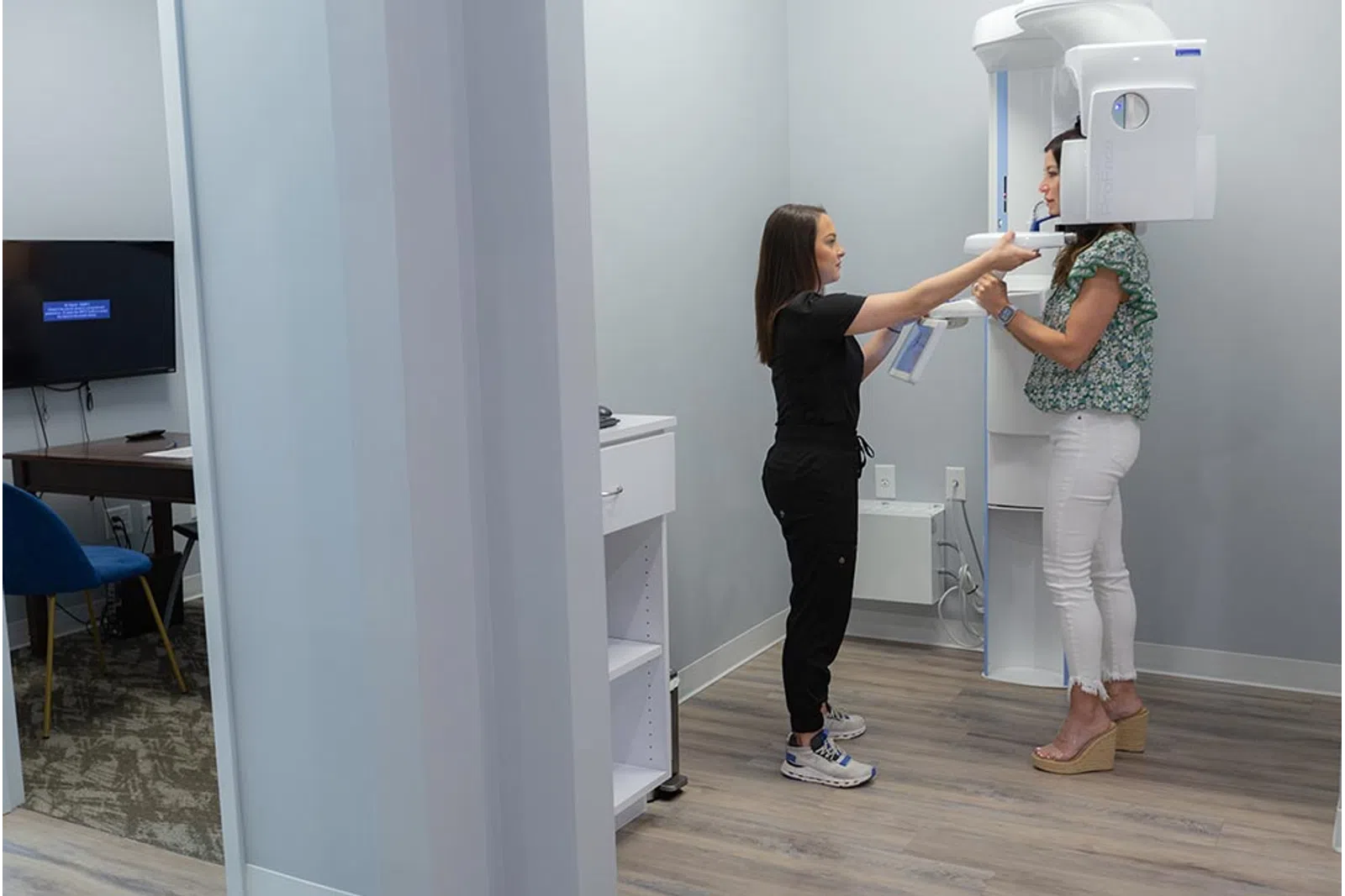 Dental technician assists patient in a floral shirt and white pants during a panoramic X-ray scan in a clinic.