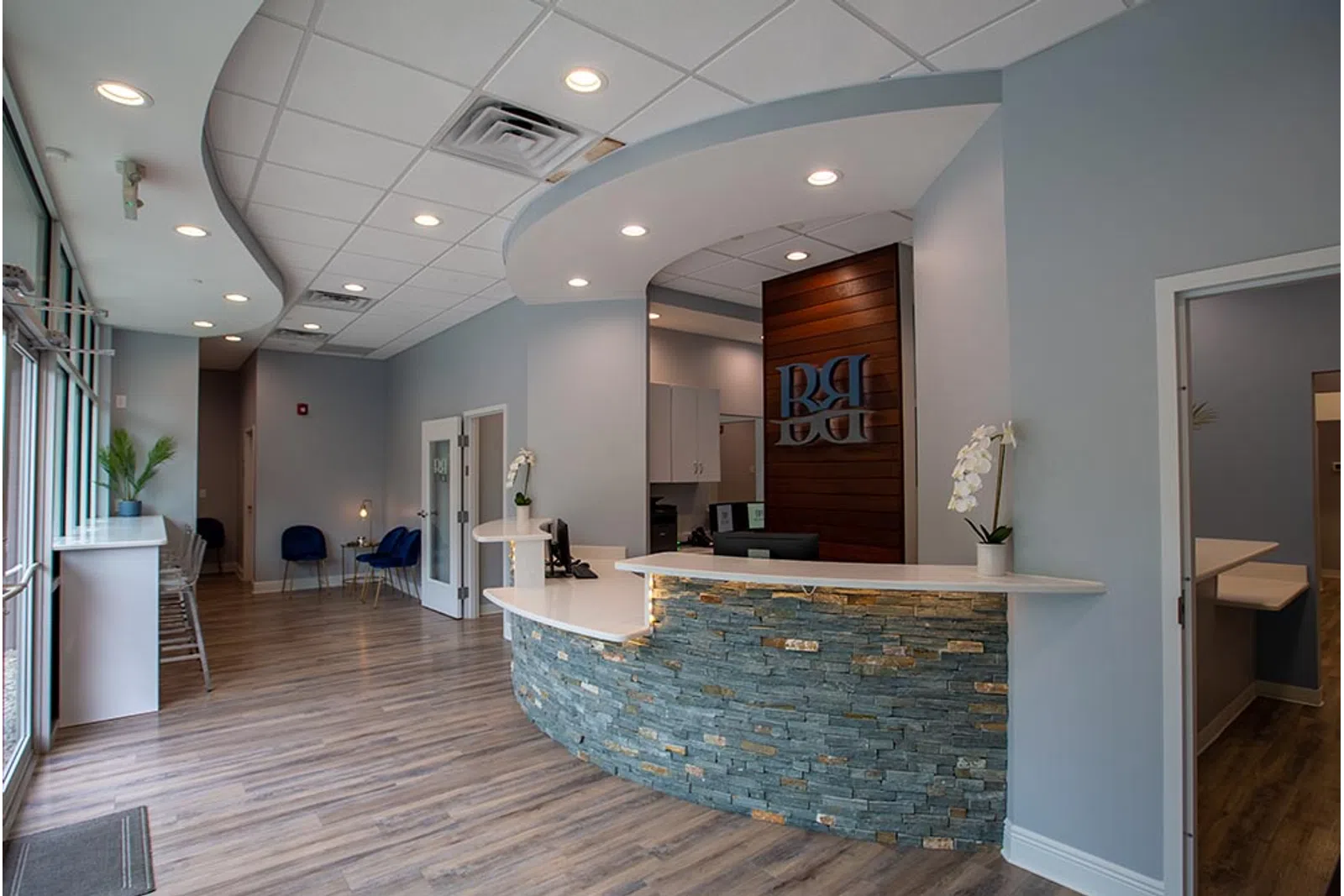 Modern dental office reception area with curved stone desk, wood flooring, blue chairs, and soft lighting.